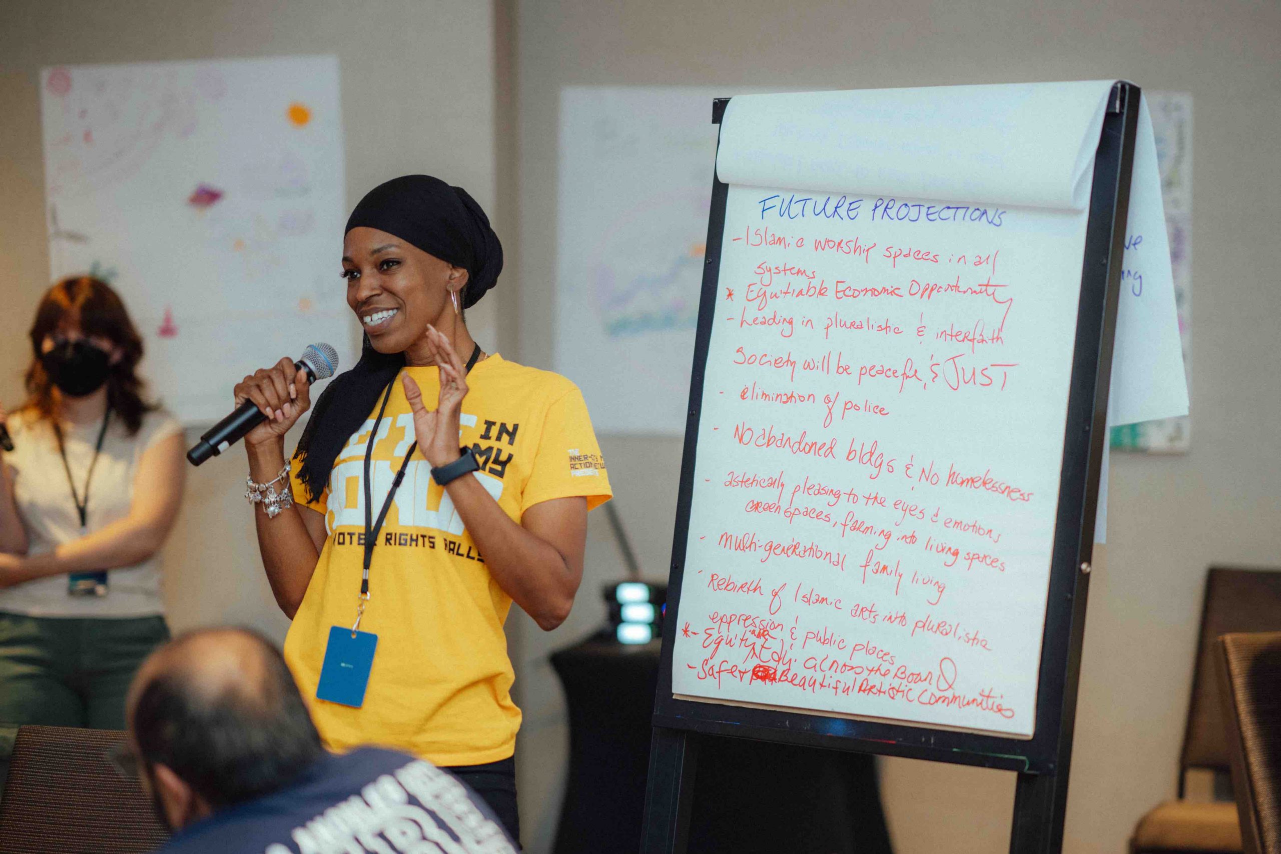 IMAN Atlanta's Kareemah Hanifa speaking into a handheld microphone next to a large easel holding a large sheet of white paper filled with handwritten notes
