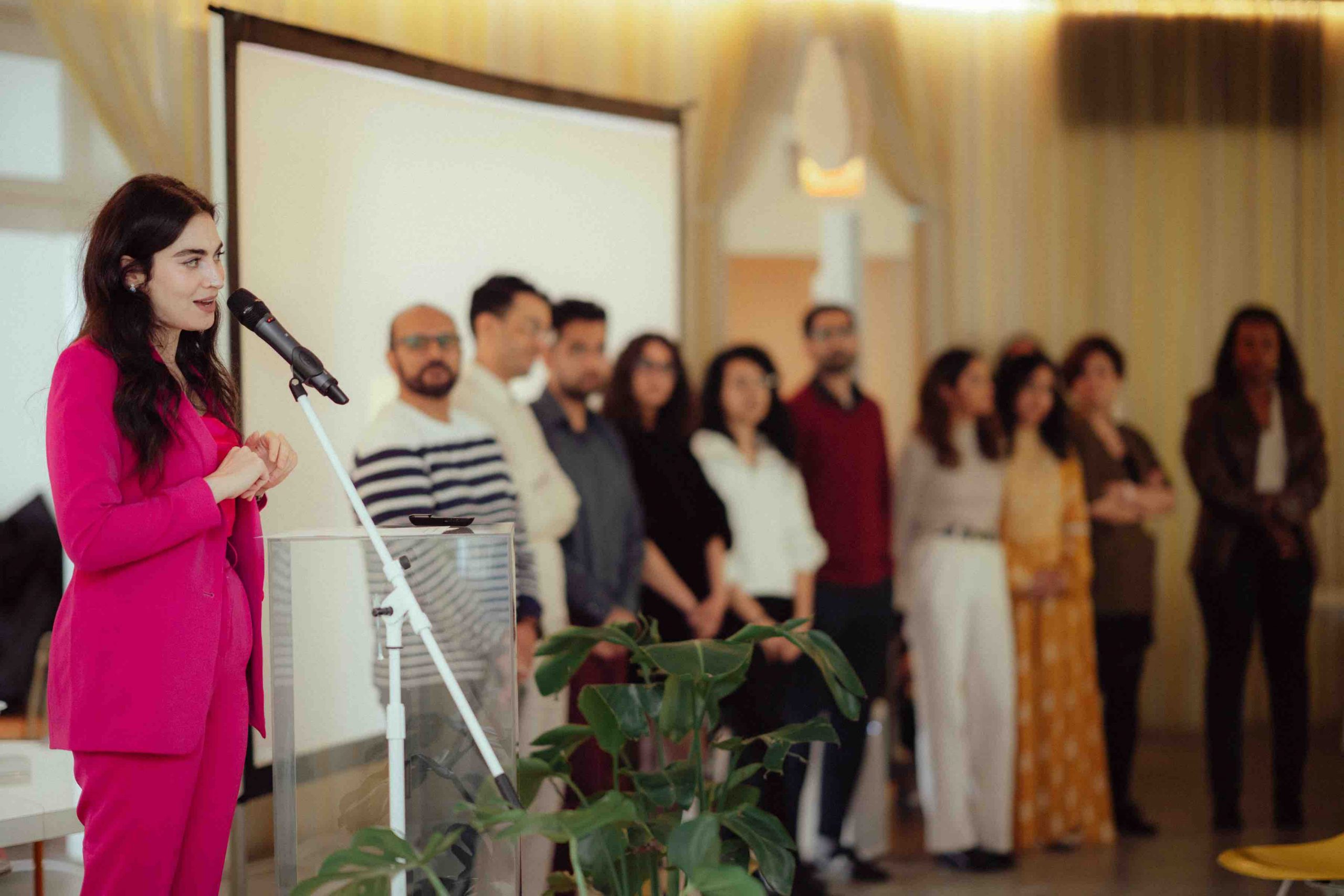 Pillars Managing Director of Culture Change Arij Mikati wearing a pink pantsuit stands in front of a clear lucite podium speaking into a microphone with a line of 10 fellows standing to her right
