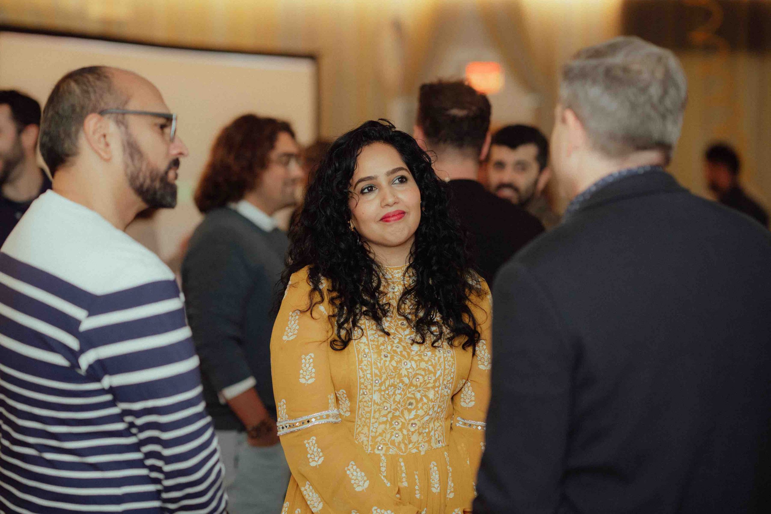 Pillars Artist Fellows Nausheen Dadabhoy and Ali Imran Zaidi speak with a reception attendee in a room full of people