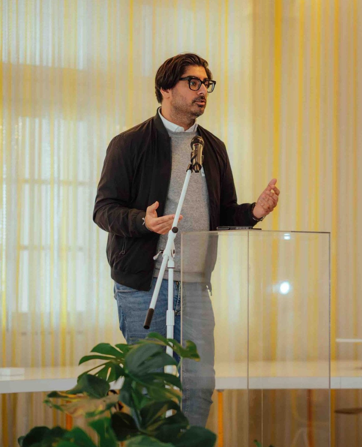 Pillars President Kashif Shaikh wearing a grey sweater, black bomber jacket, and jeans stands in front of a clear lucite podium speaking into a microphone