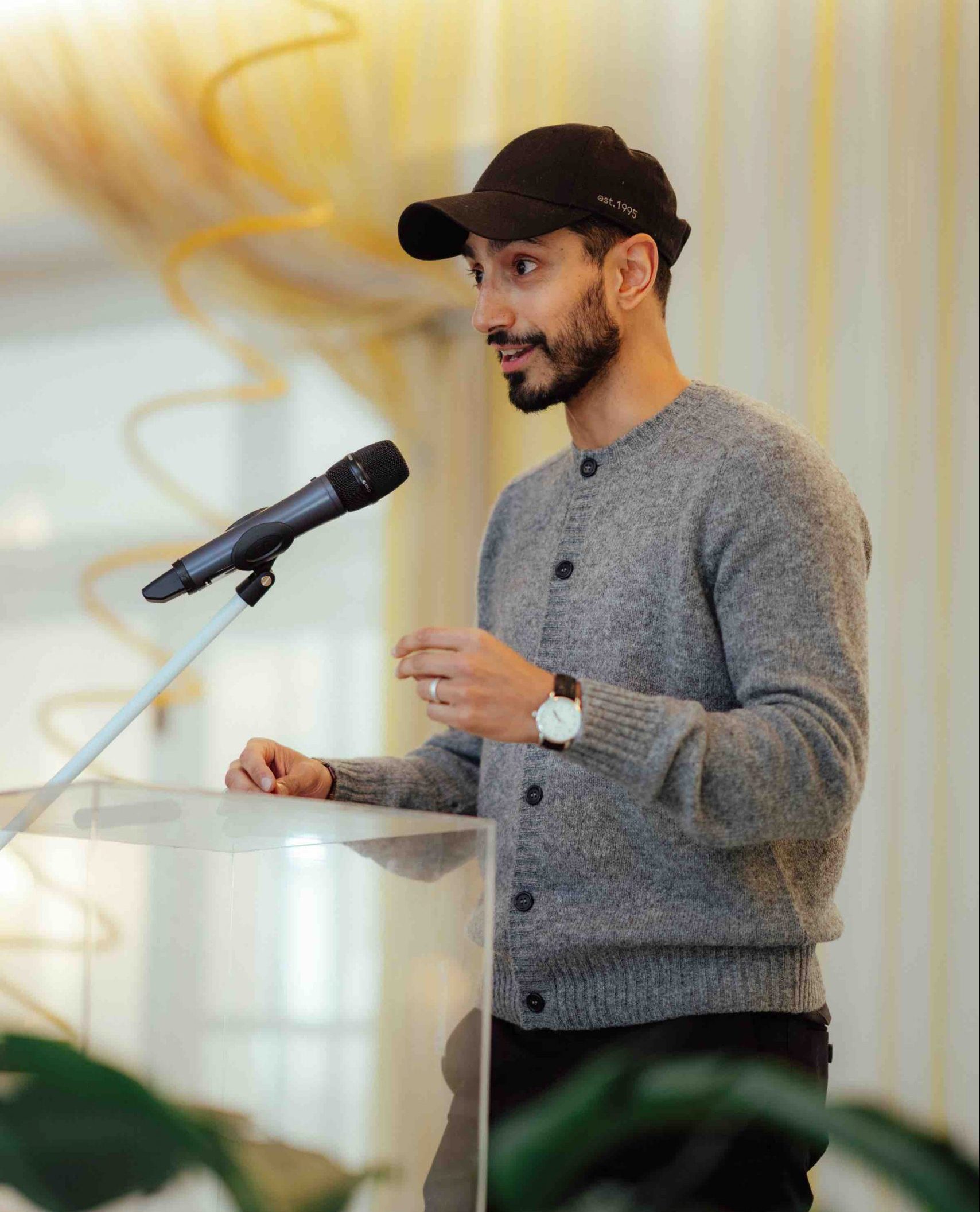 Actor Riz Ahmed wearing a grey button-up sweater and black cap stands in front of a clear lucite podium speaking into a microphone