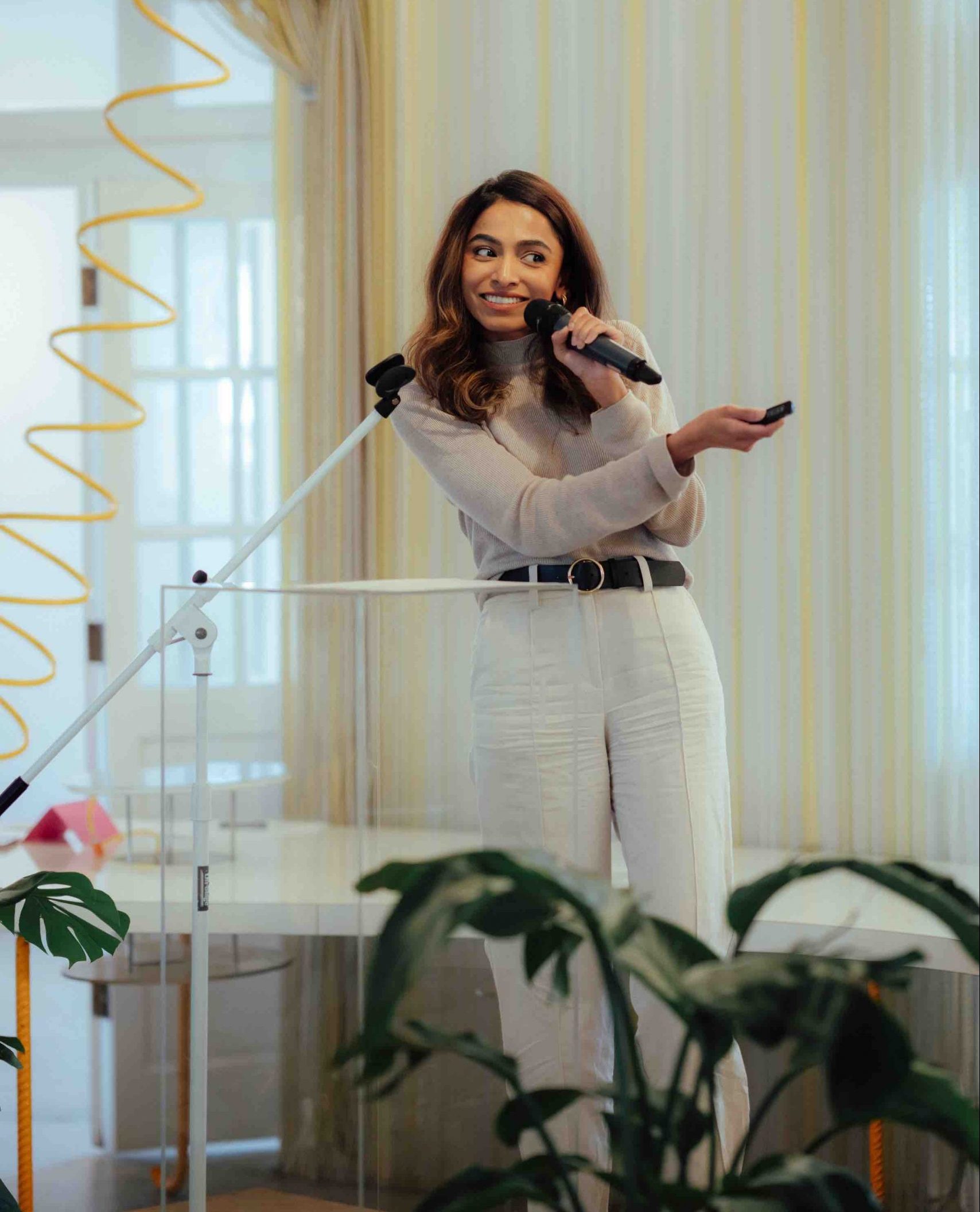 Pillars Artist Fellow Aqsa Altaf wearing a beige mock-neck sweater and linen pant smiles and holds a microphone in one hand while holding up a presentation clicker in her other hand