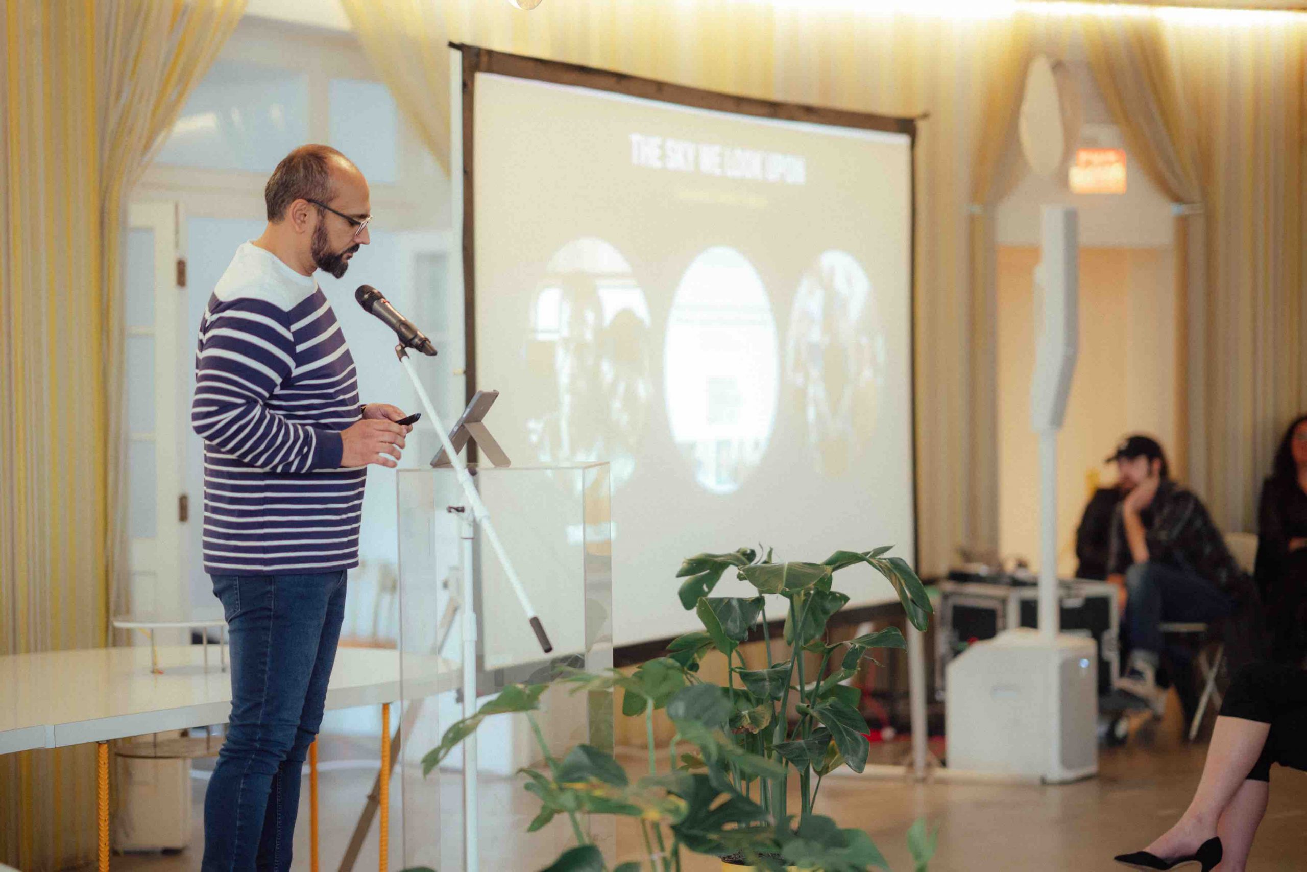 Pillars Artist Fellow Ali Imran Zaidi wearing jeans and a blue and white striped sweater stands in front of a clear lucite podium speaking into a microphone. To his right is a large projector screen