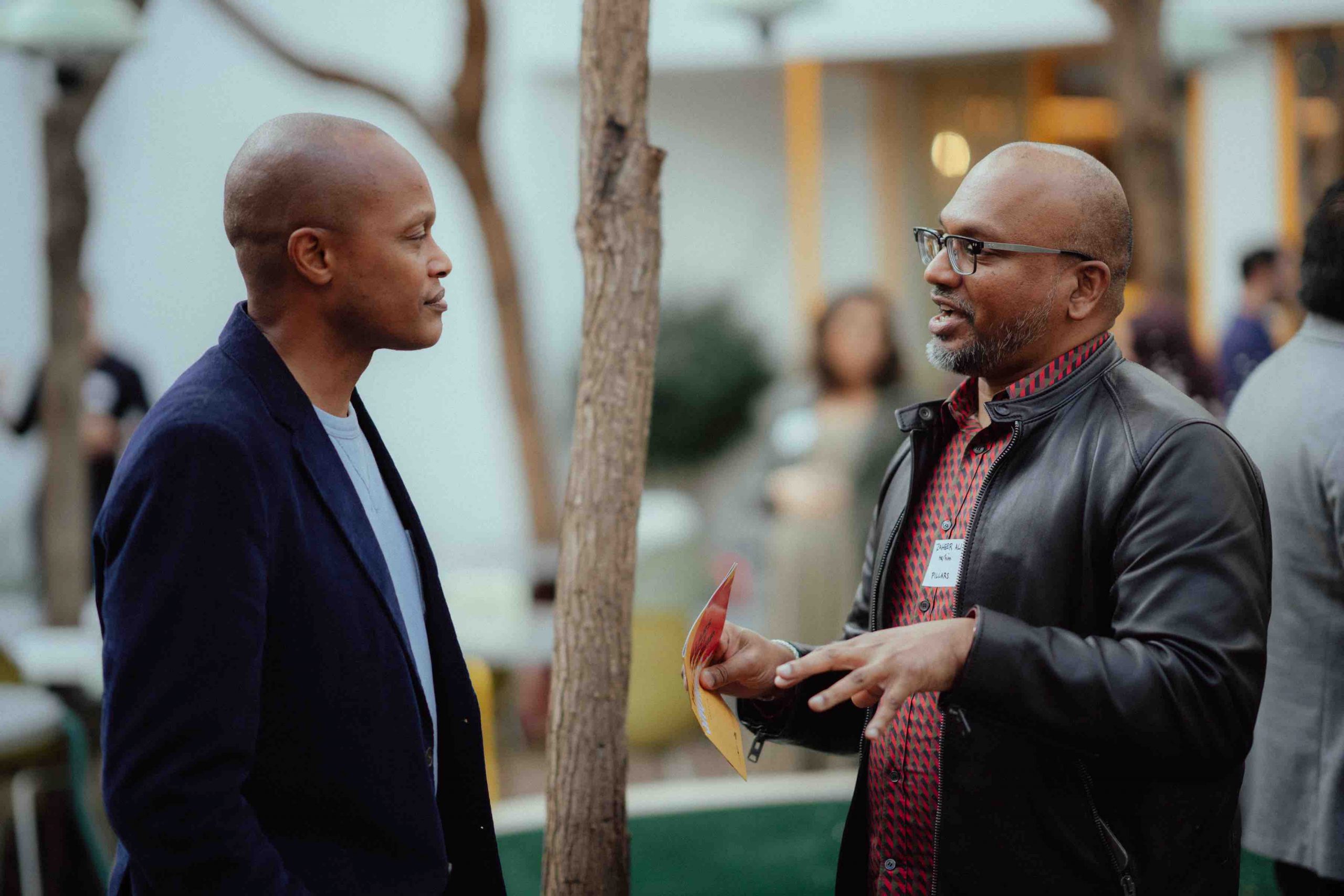 Pillars Muslim Narrative Change cohort member Zaheer Ali wearing glasses and a black leather jacket speaks with another reception attendee in an outdoor courtyard