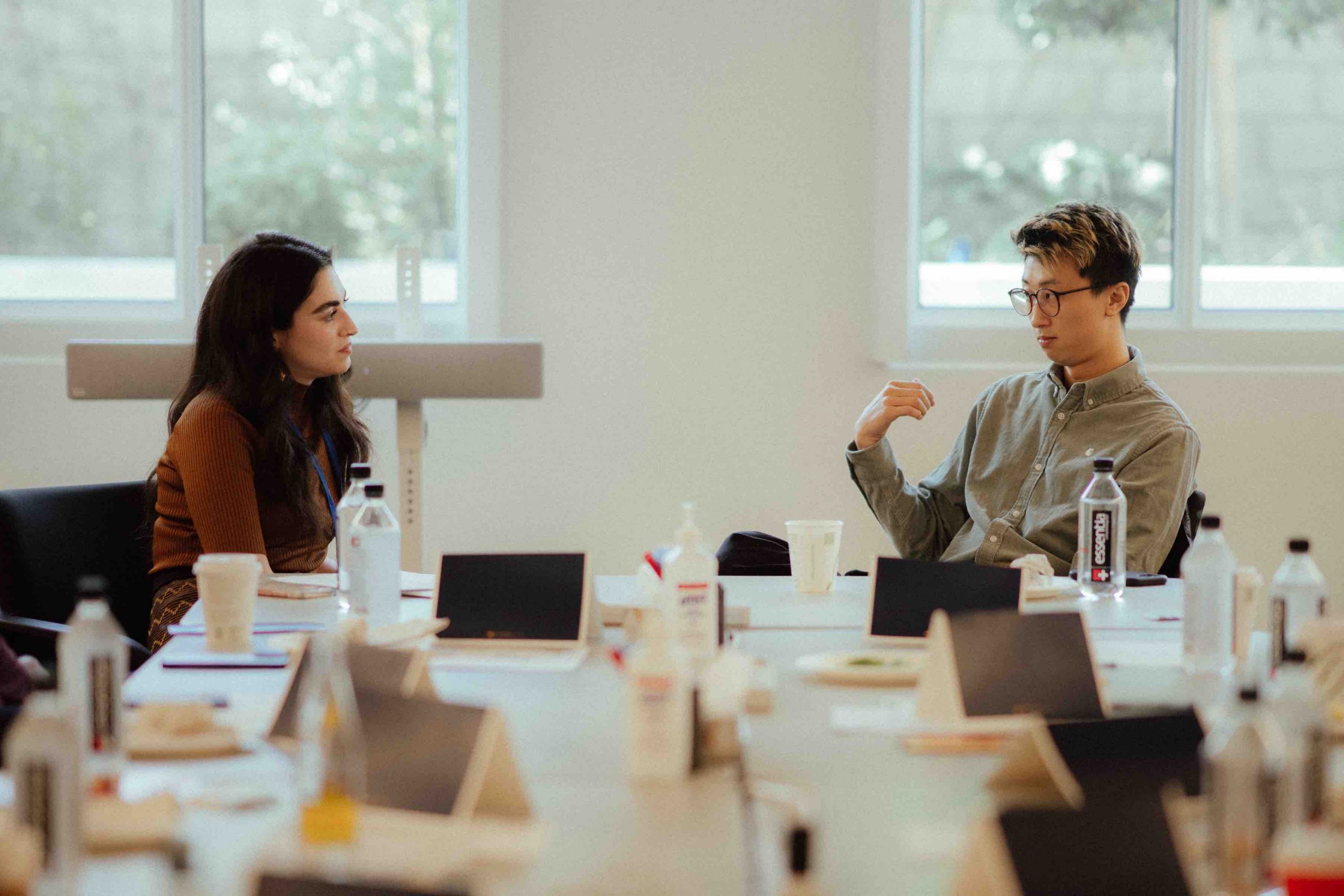 At the head of a conference table, Pillars Managing Director of Culture Change Arij Mikati looks thoughtfully at Bing Liu who has his right arm up in conversation