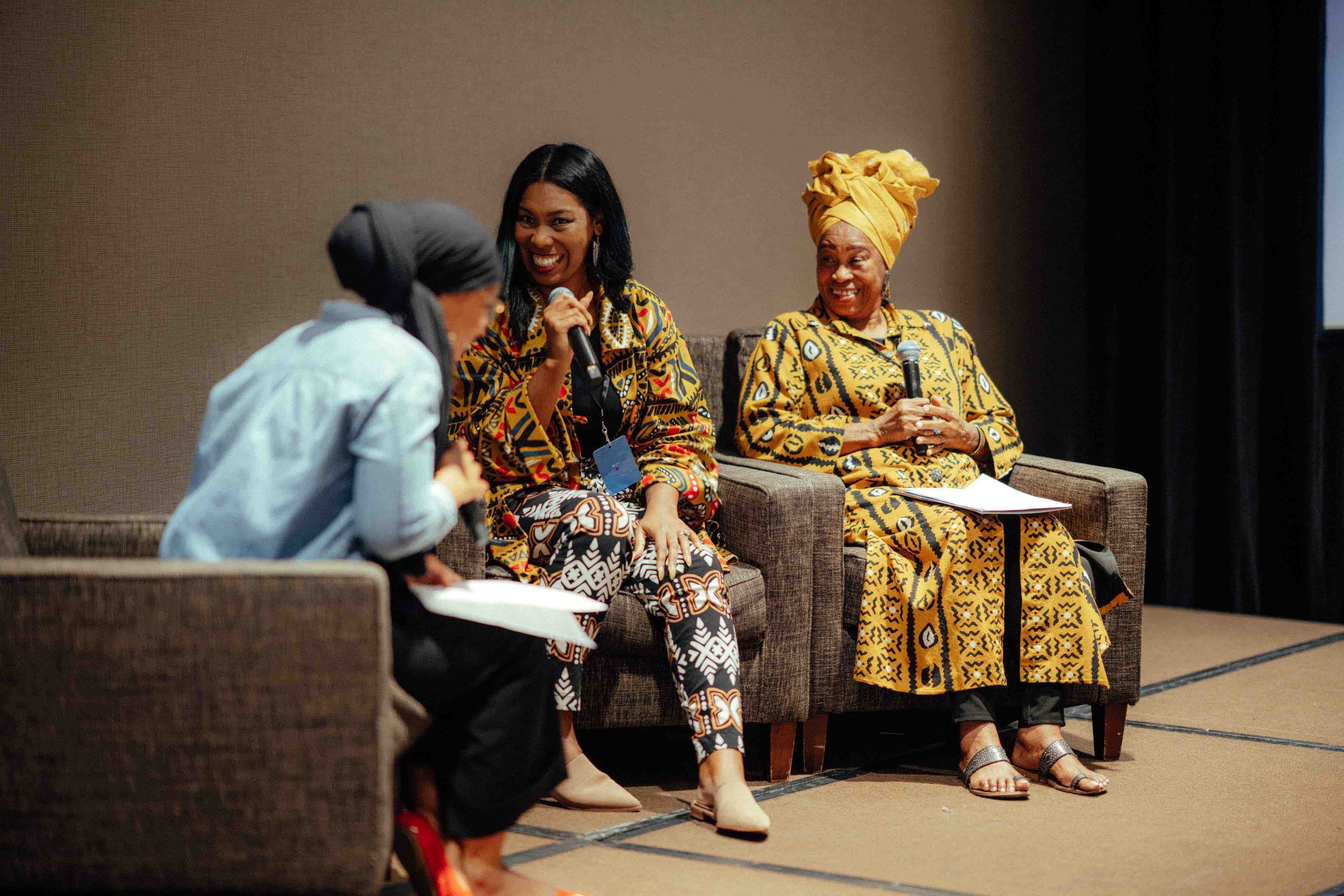 Pillars Vice President Kalia Abiade on stage with IMAN Atlanta's Aseelah Rashid and IMMC's Sister Okolo Rashid. They are all sitting in armchairs, smiling, with handheld microphones in their hands.
