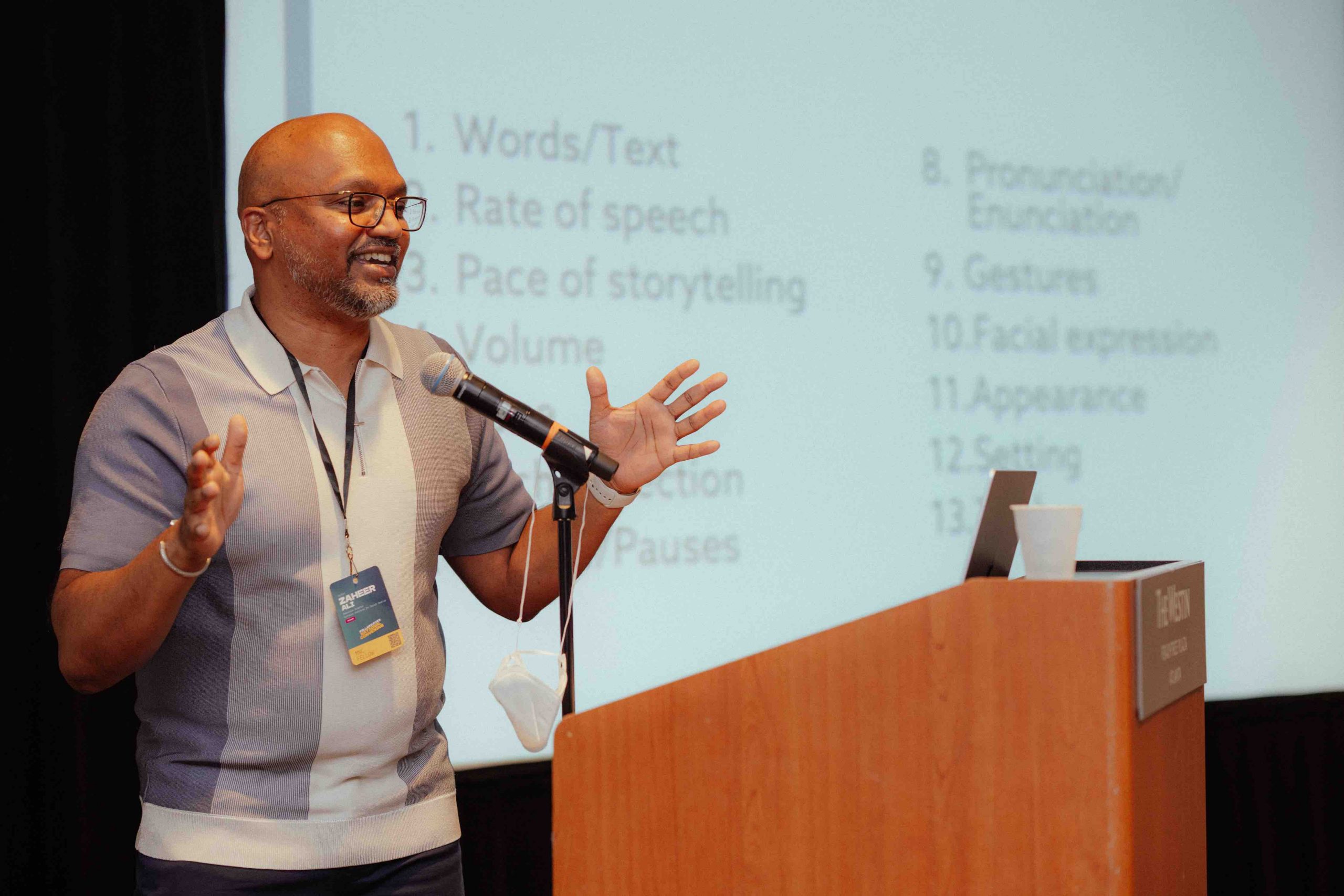 Oral historian Zaheer Ali in front of a podium speaks into a microphone, gesturing his hands animatedly