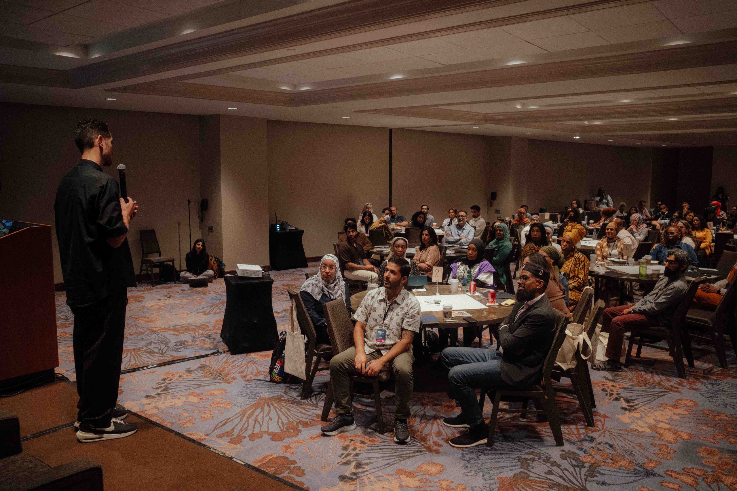 Poet Omar Offendum speaks on a stage to a handheld microphone in front of a crowd sitting around several circular tables in a conference room.