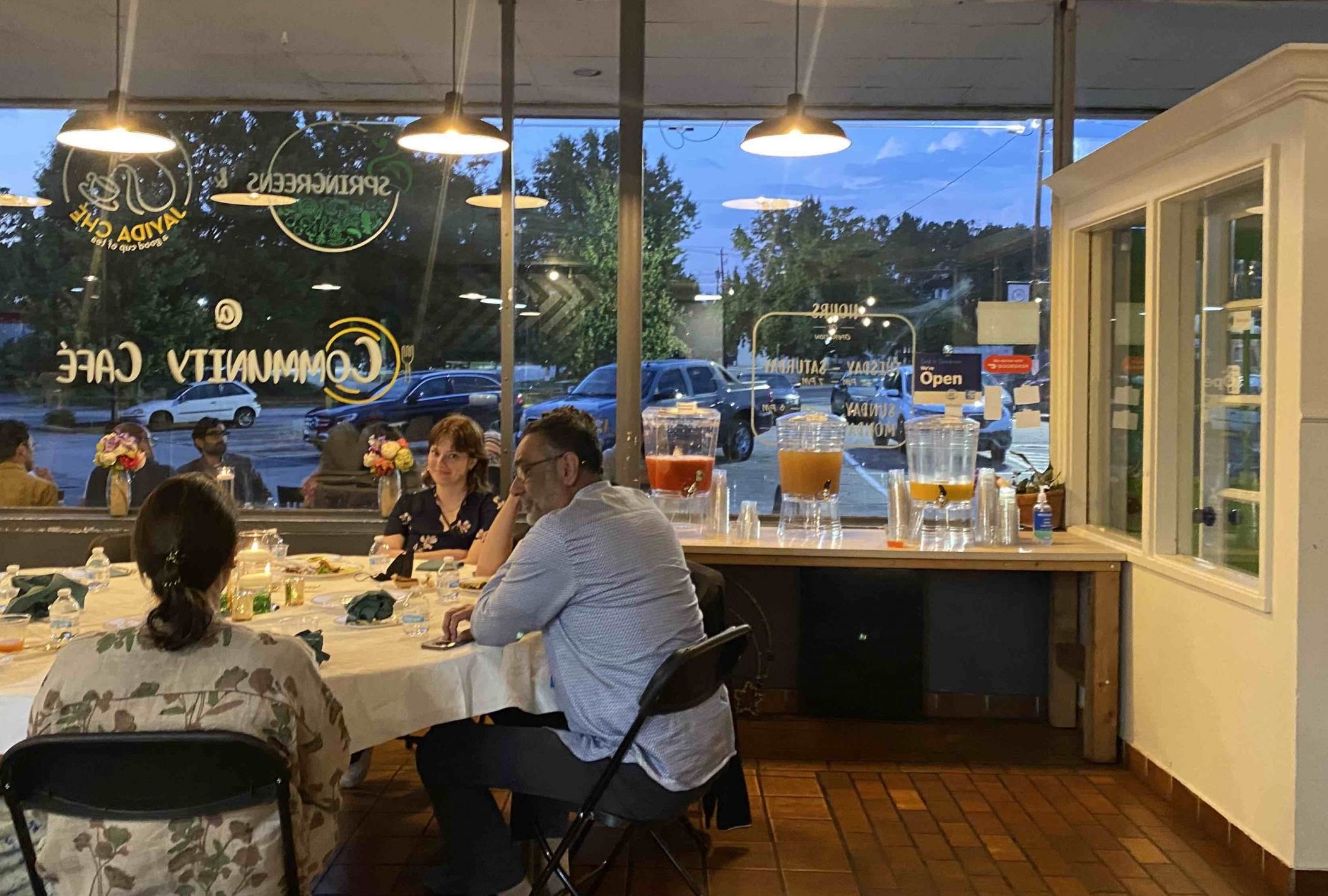 Inside Springreens cafe, Pillars National Convening attendees sit, chat, and eat around a round table, with storefront windows in the background.