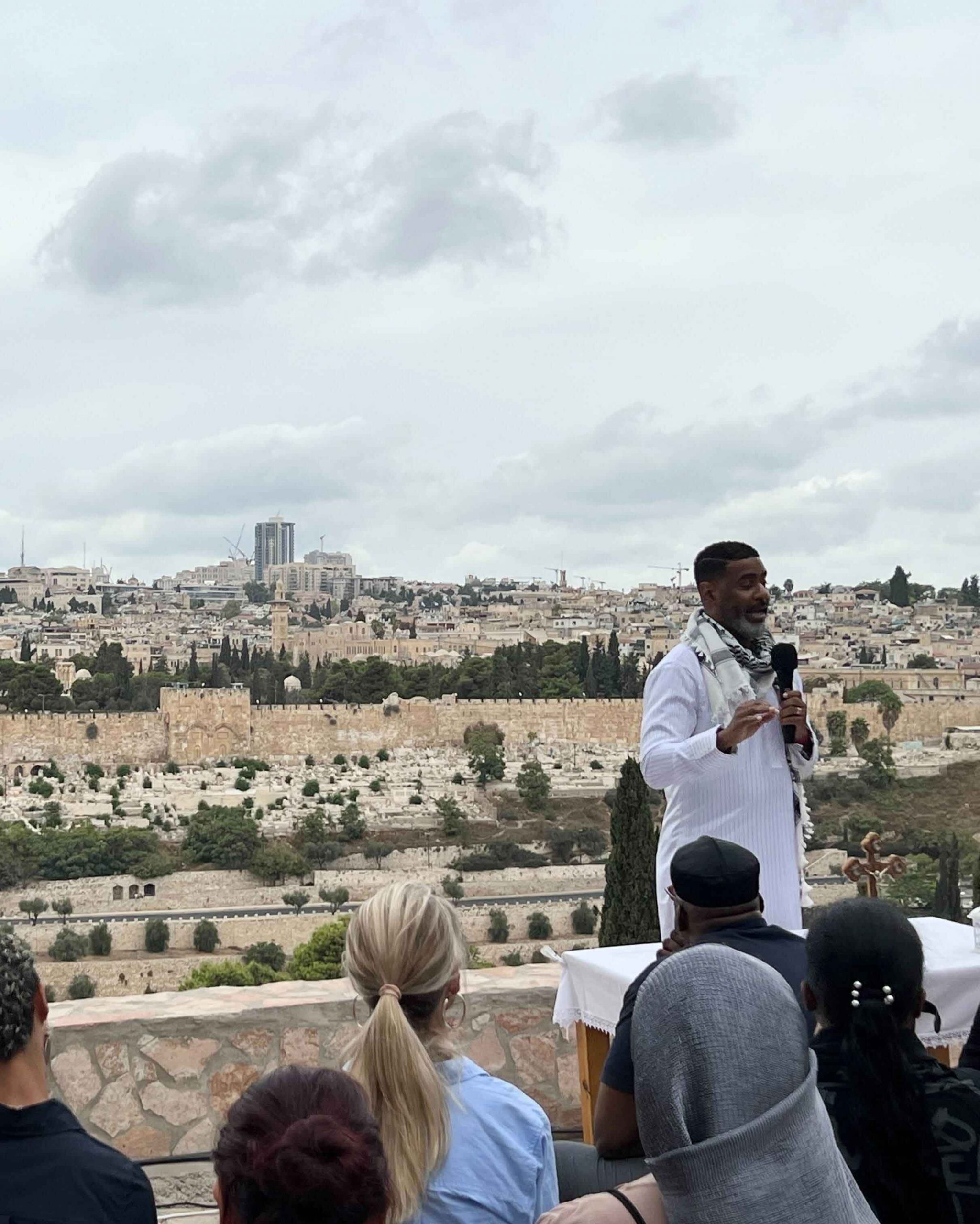 A photo of a man speaking into a microphone addresses an attentive group of people, with a view of Jerusalem in the background