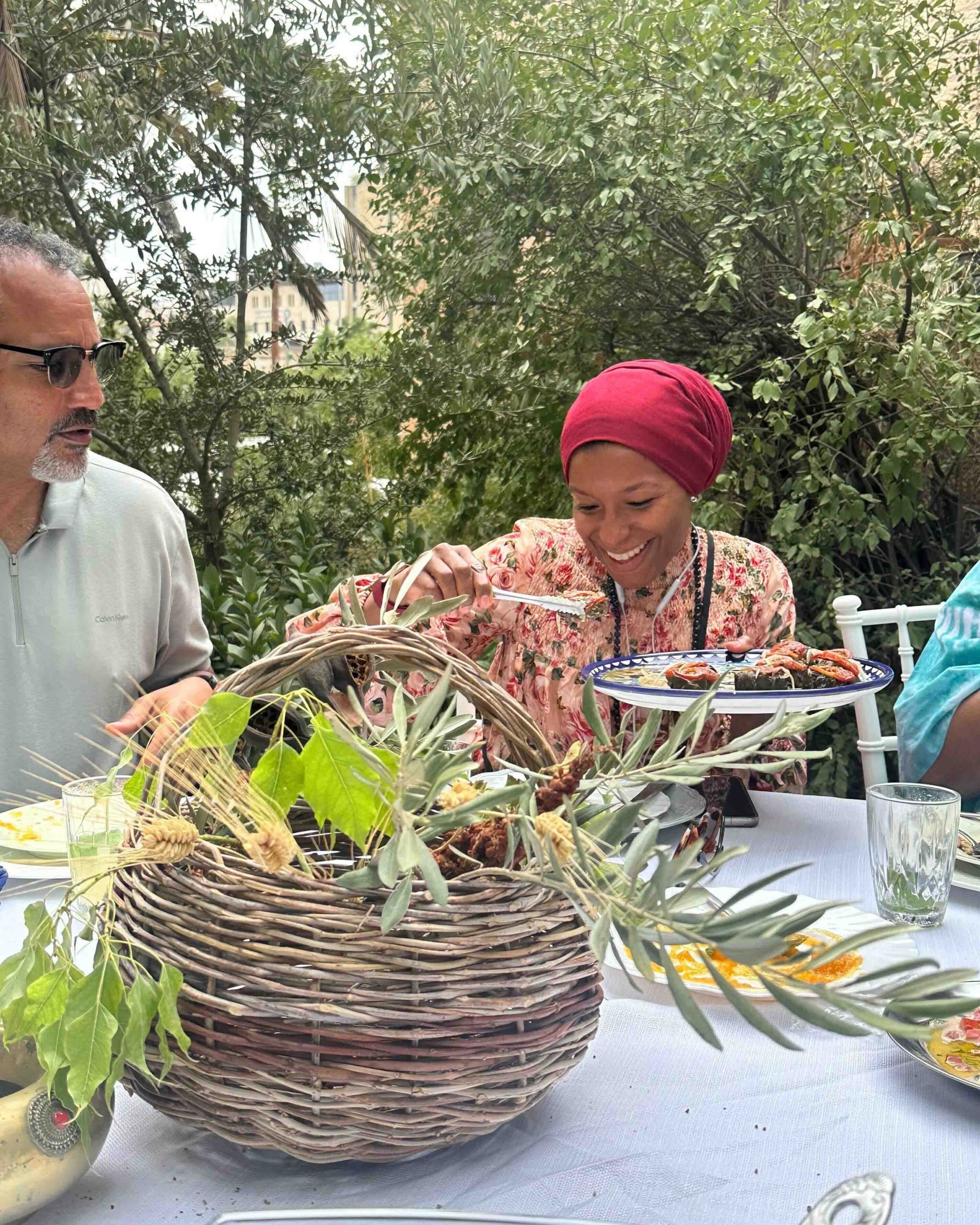 A photo of a woman sitting at a table as a part of a larger group, holding a plate and serving herself Palestinian food.