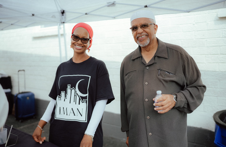 A photo of Imam Plemon El-Amin and a woman wearing a pink hijab and sunglasses smiling and standing under a white canopy outside of IMAN Atlanta’s Green ReEntry apartments